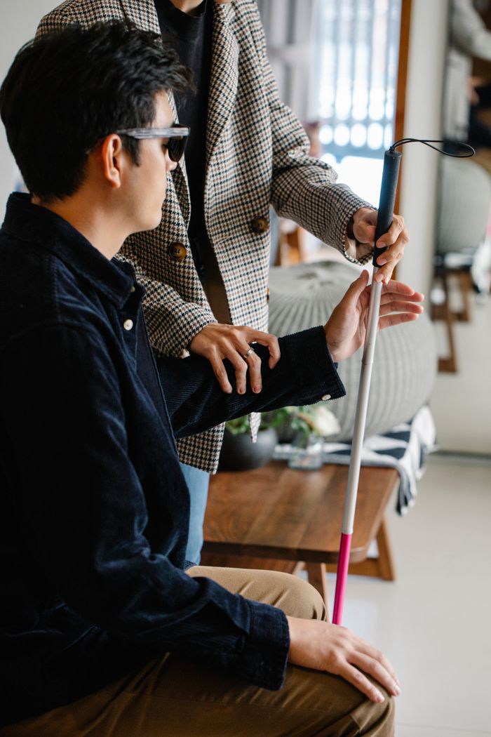 Services Blind man with sunglasses being aided by another person holding a walking stick indoors.