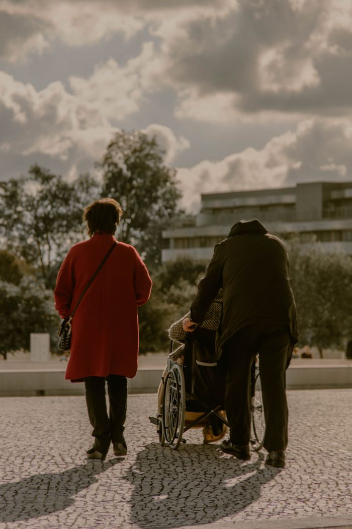 Services Two people guide a wheelchair on a sunlit pathway, embodying care and support.